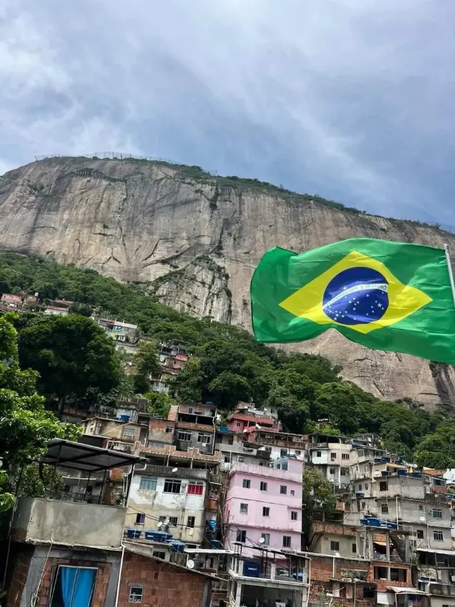 A large Brazilian flag waving against the Pedra da Gavea rock face above Rocinha