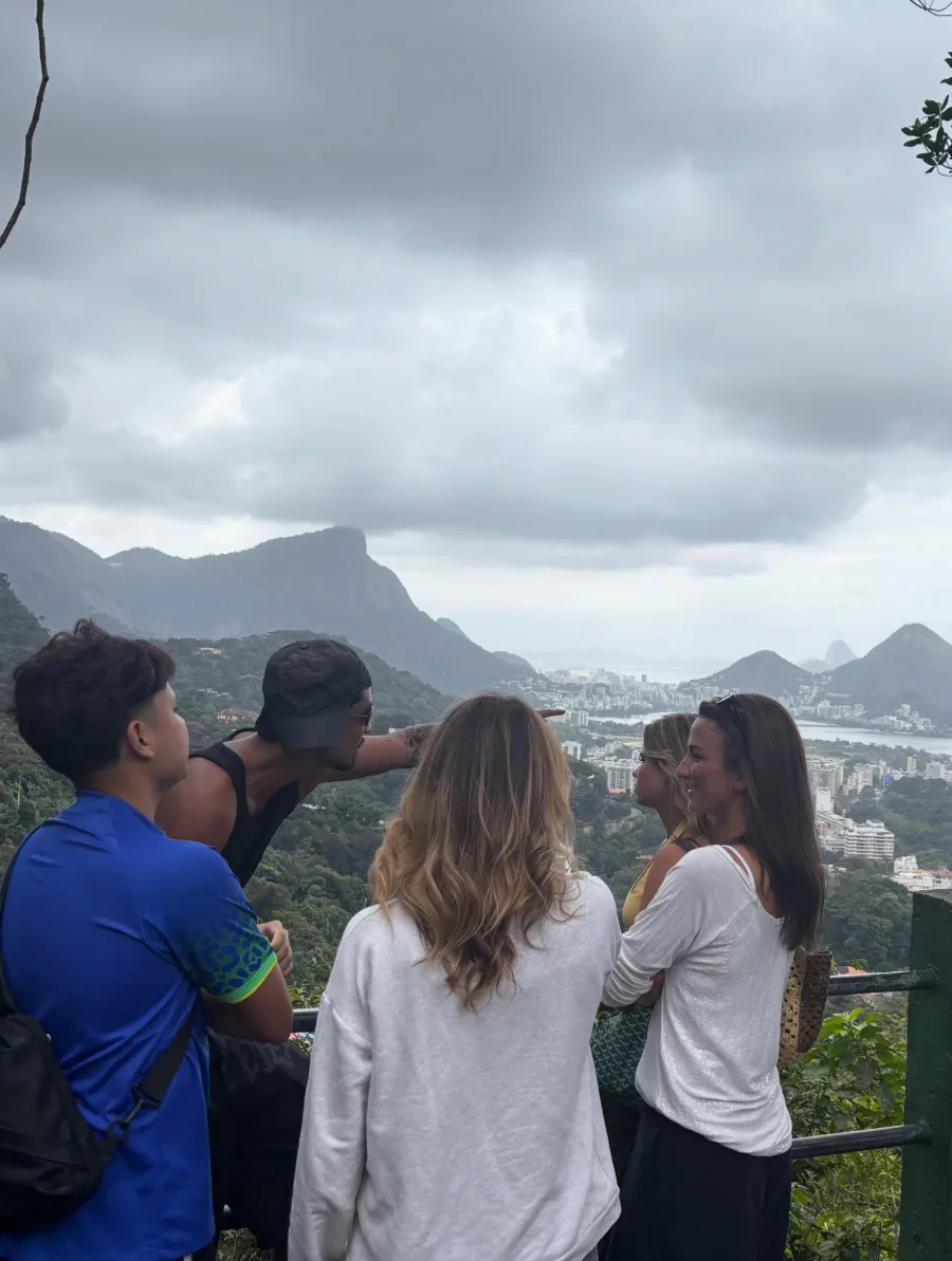 A local guide pointing out across the city from a viewpoint while four travellers follow his hand, Pedra da Gavea and the coastline under heavy cloud