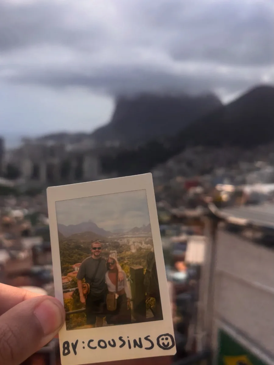 A polaroid of two travellers held up against the same view it was taken in, with BY: COUSINS written on the white border